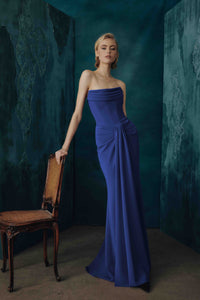 A woman models a strapless royal blue floor-length gown in a moody studio setting with dark teal textured walls and a wooden chair beside her.