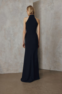A woman stands with her back to the camera wearing a sleeveless, high-neck floor-length gown in a very dark navy or near-black fabric, posed against a textured beige studio wall and floor.