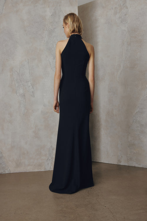 A woman stands with her back to the camera wearing a sleeveless, high-neck floor-length gown in a very dark navy or near-black fabric, posed against a textured beige studio wall and floor.