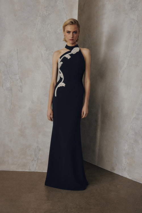 A woman stands in a studio wearing a floor-length navy gown with a high neckline and a large white abstract floral or leaf-like applique running diagonally across the bodice and down one side. The background consists of textured neutral walls in beige and gray tones.