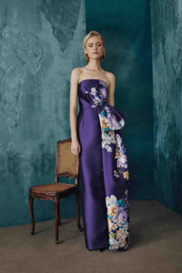 A woman models a strapless floor-length gown in deep purple satin with a large floral print panel on the skirt, standing in a studio with a textured teal wall and a wooden chair.