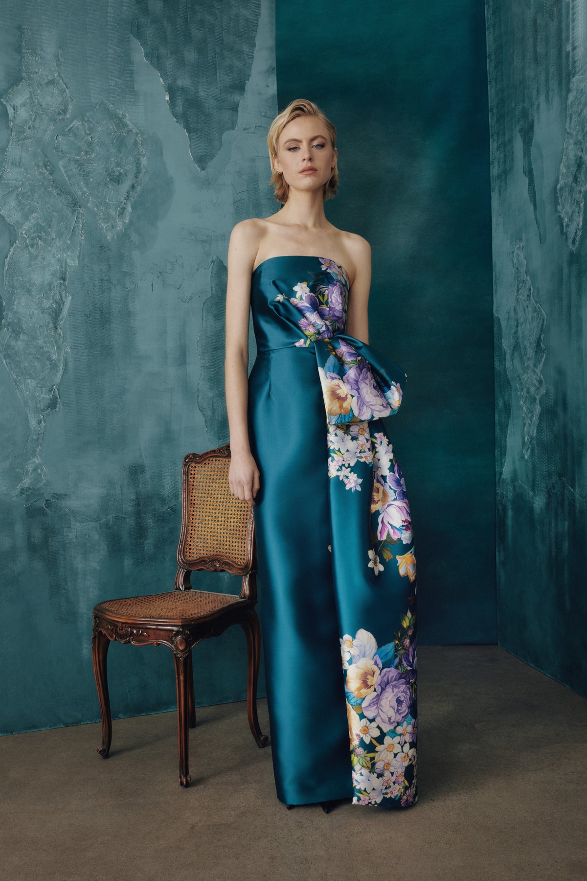 A woman models a strapless teal satin gown with a large floral print draped over one side, standing in a studio with textured teal walls and a wooden chair.