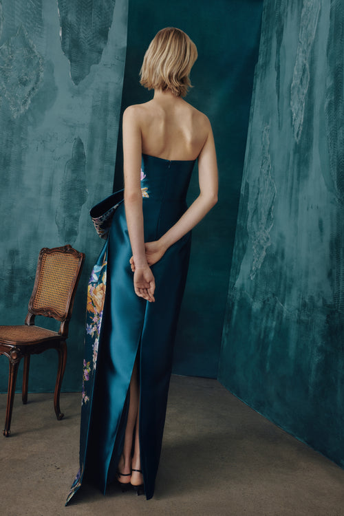 A woman stands with her back to the camera wearing a strapless teal satin gown with floral embroidery and a high slit, posed in a moody room with textured teal walls and a wooden chair.