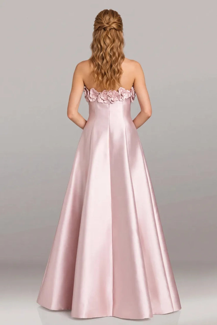 A woman viewed from the back wearing a strapless formal gown in light blush pink satin with floral embellishments across the upper back, against a plain light gray studio background.