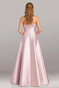 A woman viewed from the back wearing a strapless formal gown in light blush pink satin with floral embellishments across the upper back, against a plain light gray studio background.