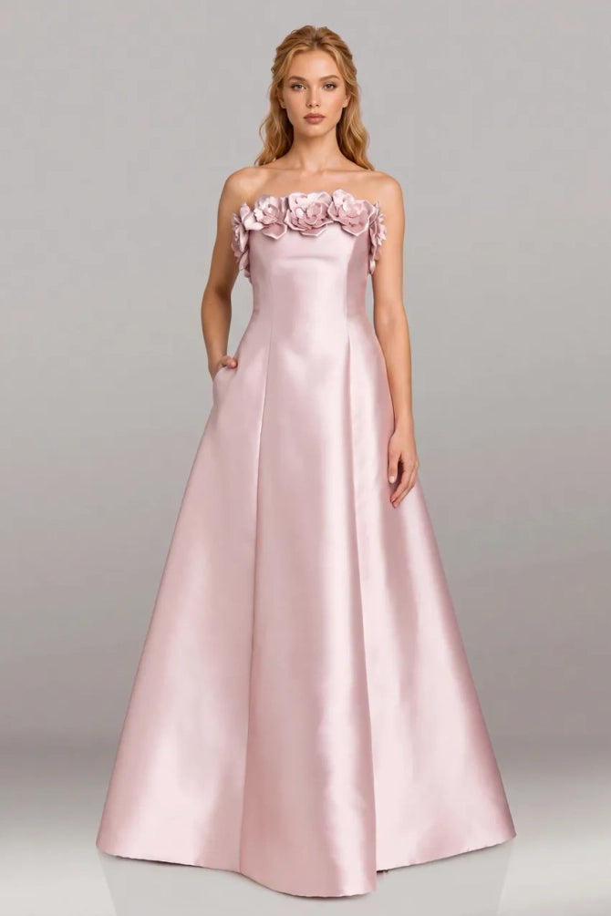 A woman modeling a floor-length, strapless A-line gown in light blush pink satin, featuring a ruffled floral neckline, against a neutral gray studio background.