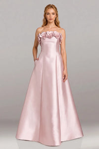 A woman modeling a floor-length, strapless A-line gown in light blush pink satin, featuring a ruffled floral neckline, against a neutral gray studio background.