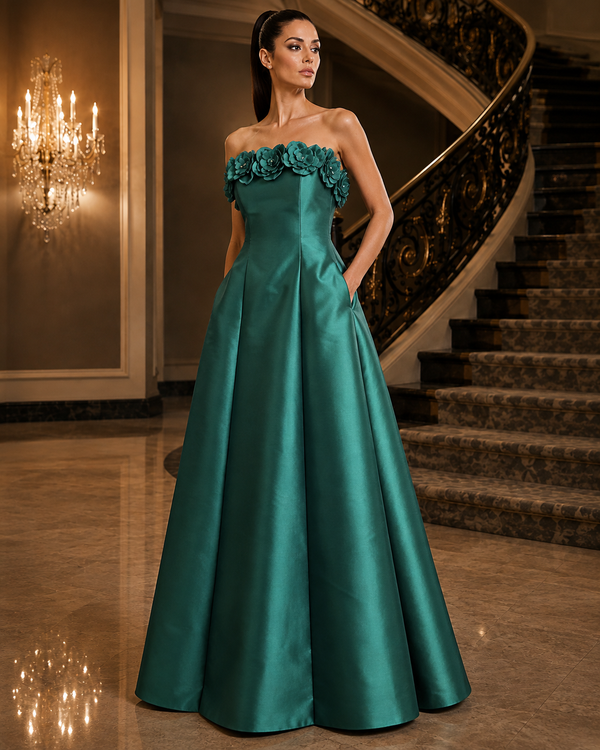 A woman models a strapless floor-length gown in a grand interior with a staircase and chandelier. The dress is a rich teal green satin with 3D floral applique across the neckline.