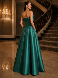 A woman wearing a strapless emerald green satin ball gown with a ruffled sweetheart bodice, photographed from the back in an elegant indoor setting with a staircase and chandelier.