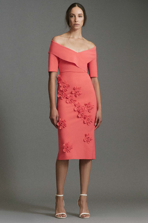 Woman wearing a coral off-shoulder dress with floral embellishments on a gray background