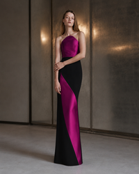 A woman models a floor-length satin gown with a color-blocked design in black and vivid magenta. The dress features a one-shoulder neckline and a fitted silhouette, shown against a modern textured gray wall with warm lighting.