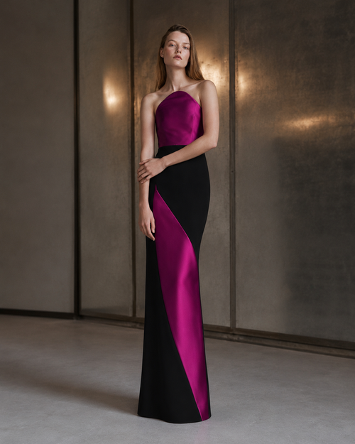 A woman models a floor-length satin gown with a color-blocked design in black and vivid magenta. The dress features a one-shoulder neckline and a fitted silhouette, shown against a modern textured gray wall with warm lighting.