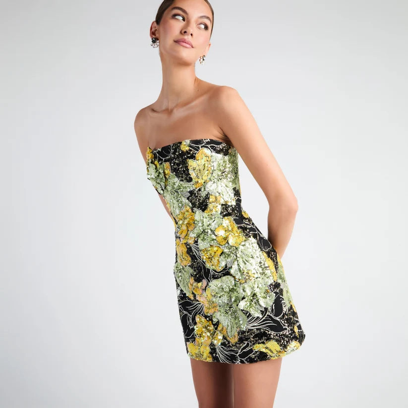 A woman modeling a strapless mini dress with a black base and a vibrant yellow, green, and white floral pattern, photographed against a solid light gray studio background.