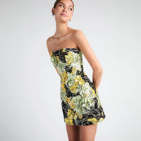 A woman modeling a strapless mini dress with a black base and a vibrant yellow, green, and white floral pattern, photographed against a solid light gray studio background.