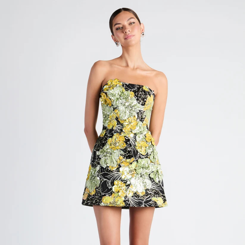 A woman modeling a strapless mini dress with a black base and a vibrant yellow, green, and white floral pattern, set against a plain light gray studio background.