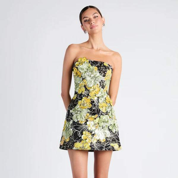 A woman modeling a strapless mini dress with a black base and a vibrant yellow, green, and white floral pattern, set against a plain light gray studio background.