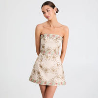 A woman models a strapless tube dress with a flared skirt. The dress has a warm beige base with an all-over embroidered pattern of gold florals and small pink and green leaves.