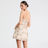 A woman modeling a strapless mini dress from the back, featuring a beige base with a gold floral pattern and subtle pink accents, set against a plain light gray background.
