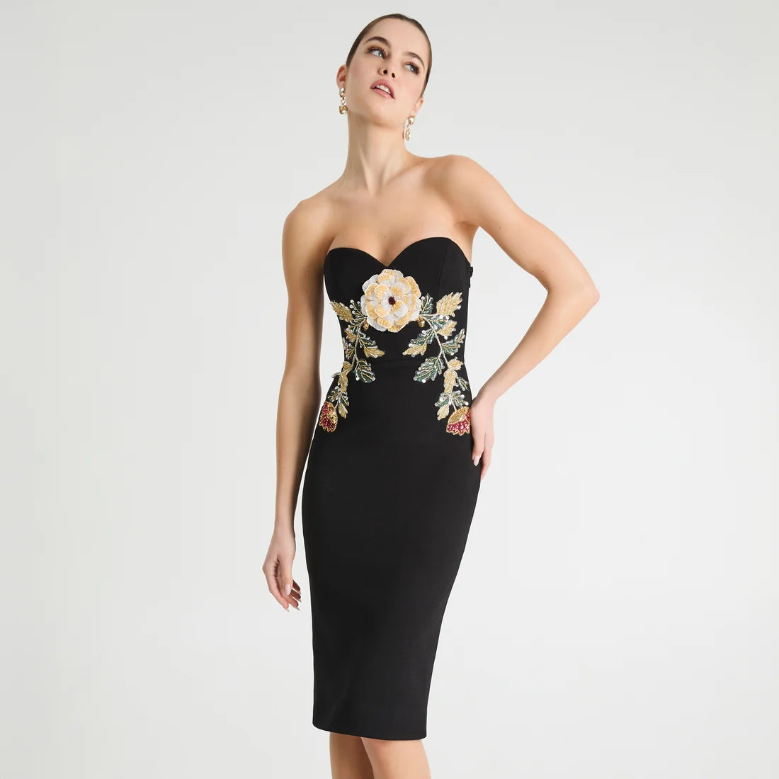 A woman models a strapless black bodycon dress with a sweetheart neckline and a large floral embroidery design on the bodice and hips. The embroidery features cream, gold, and green tones.