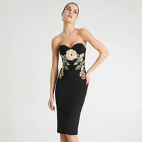 A woman models a strapless black bodycon dress with a sweetheart neckline and a large floral embroidery design on the bodice and hips. The embroidery features cream, gold, and green tones.