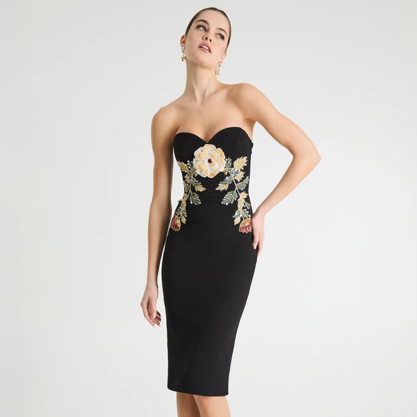 A woman models a strapless black bodycon dress with a sweetheart neckline and a large floral embroidery design on the bodice and hips. The embroidery features cream, gold, and green tones.