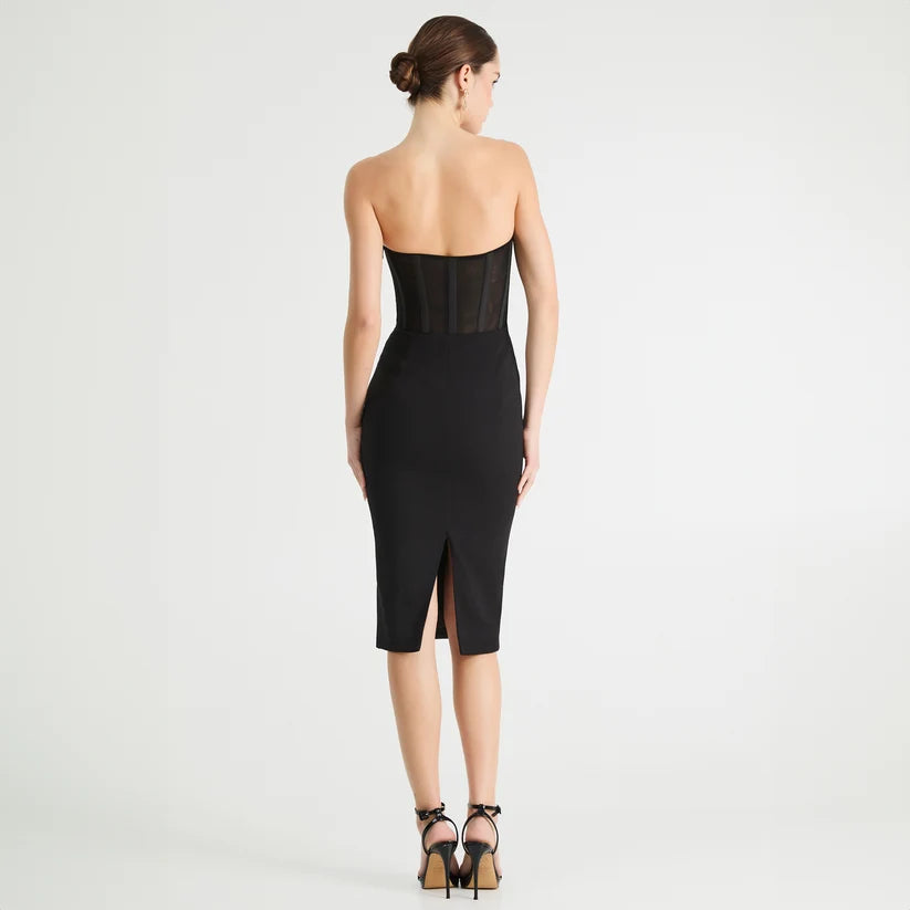 A woman stands with her back to the camera wearing a strapless black midi dress with sheer mesh panels at the waist and a front slit, paired with black high-heeled sandals against a light gray studio background.