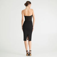 A woman stands with her back to the camera wearing a strapless black midi dress with sheer mesh panels at the waist and a front slit, paired with black high-heeled sandals against a light gray studio background.