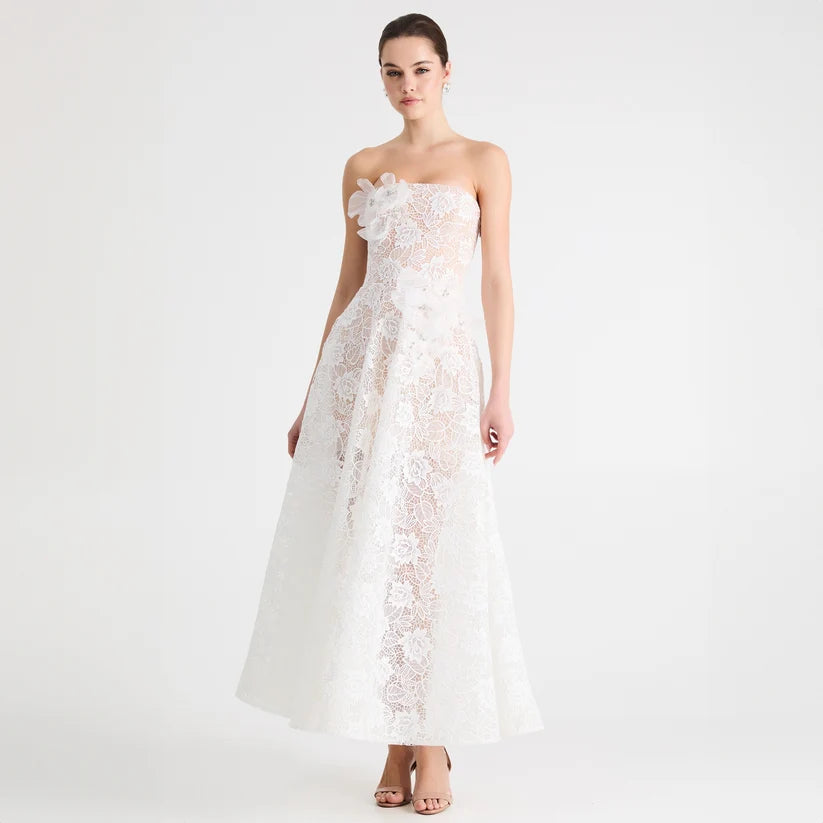 A woman modeling a strapless white lace gown with a large floral applique at the bust and a full floor-length skirt, photographed against a clean white studio background.