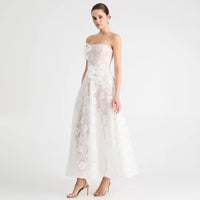 A woman modeling a strapless white lace gown with a fitted bodice, one-shoulder floral detail, and a full floor-length skirt, photographed against a clean white studio background.