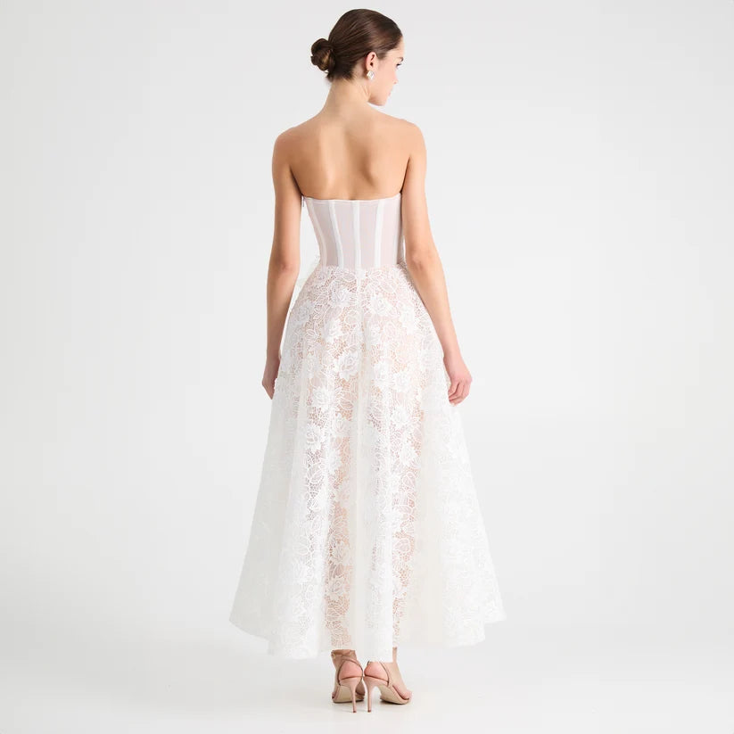 A woman shown from the back wearing a strapless white lace gown with a structured corset-style bodice and a full tulle skirt, photographed against a clean light gray studio background.