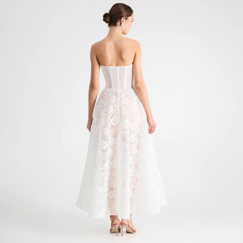 A woman shown from the back wearing a strapless white lace gown with a structured corset-style bodice and a full tulle skirt, photographed against a clean light gray studio background.