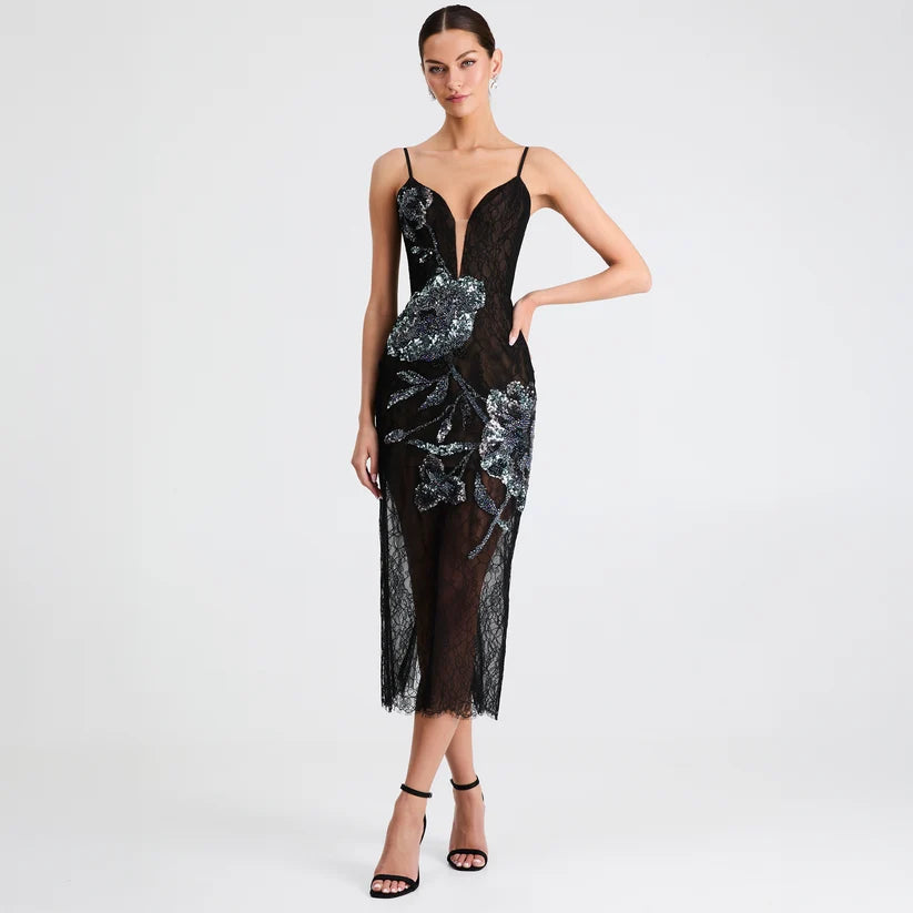 A model wears a black lace midi dress with silver sequined floral patterns, featuring spaghetti straps, a plunging V-neckline, and a sheer overlay, set against a plain light gray studio background.