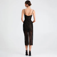 A woman viewed from the back wearing a black lace spaghetti-strap dress with a low open back and sheer mesh overlay, paired with black ankle-strap heels, against a light gray studio background.
