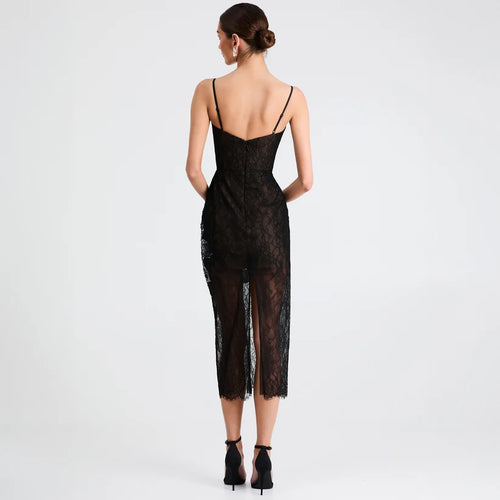 A woman viewed from the back wearing a black lace spaghetti-strap dress with a low open back and sheer mesh overlay, paired with black ankle-strap heels, against a light gray studio background.