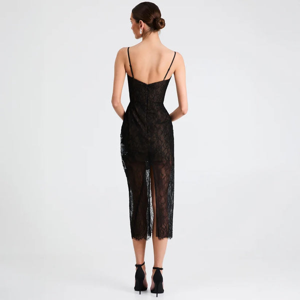 A woman viewed from the back wearing a black lace spaghetti-strap dress with a low open back and sheer mesh overlay, paired with black ankle-strap heels, against a light gray studio background.