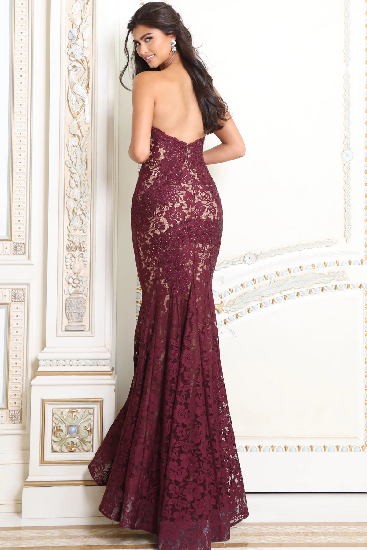 A woman modeling a strapless burgundy lace mermaid gown with an open back, standing in front of an ornate cream-colored wall.