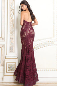 A woman modeling a strapless burgundy lace mermaid gown with an open back, standing in front of an ornate cream-colored wall.