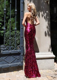 A woman stands outdoors in front of a stone wall and black metal gate, wearing a floor-length, backless sequined gown in a vivid deep pink or magenta tone. The photo is taken from behind to show the full silhouette of the dress.