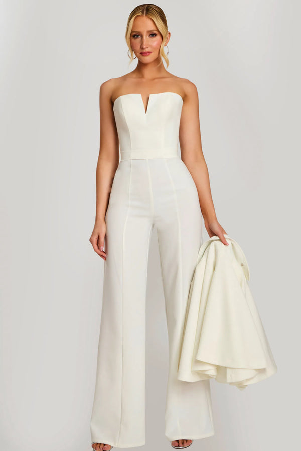 Woman wearing a strapless white jumpsuit holding a matching jacket against a plain background