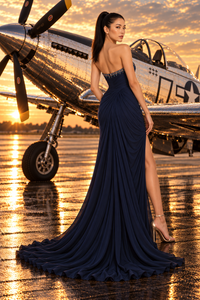 A woman stands on a wet tarmac in front of a vintage military airplane at sunset, wearing a strapless navy blue evening gown with a high slit and long train.
