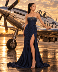A woman stands on a wet tarmac in front of a vintage silver airplane at sunset, wearing a strapless navy blue evening gown with a high thigh slit and embellished bodice.