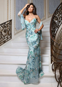 A woman models a floor-length, off-the-shoulder formal gown with a sweetheart neckline and a fitted mermaid silhouette, featuring a light blue and white floral pattern, posed on an elegant white staircase with ornate metal railings.