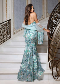A woman wearing a formal off-the-shoulder gown with a floral print in shades of teal, mint green, and light blue, standing on a white staircase next to an ornate metal railing.