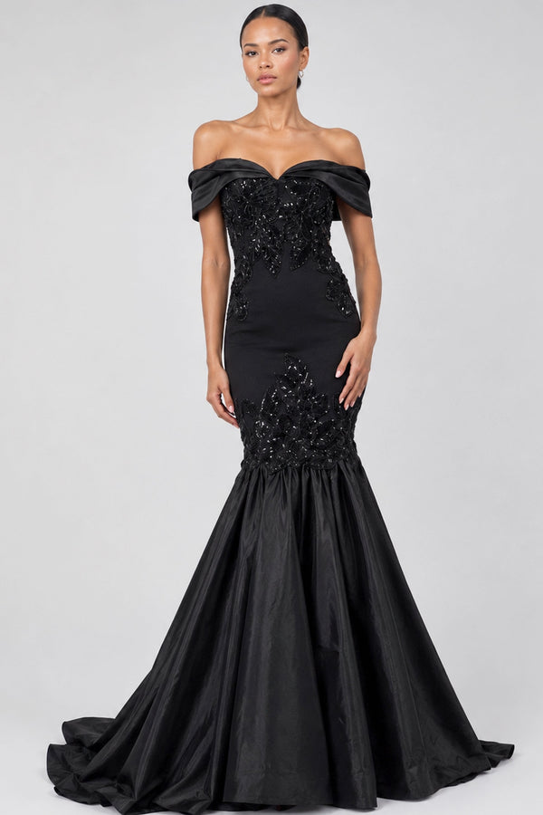 A woman modeling an off-the-shoulder black mermaid gown with a fitted bodice, beaded embellishments, and a flowing satin skirt against a light gray studio background.