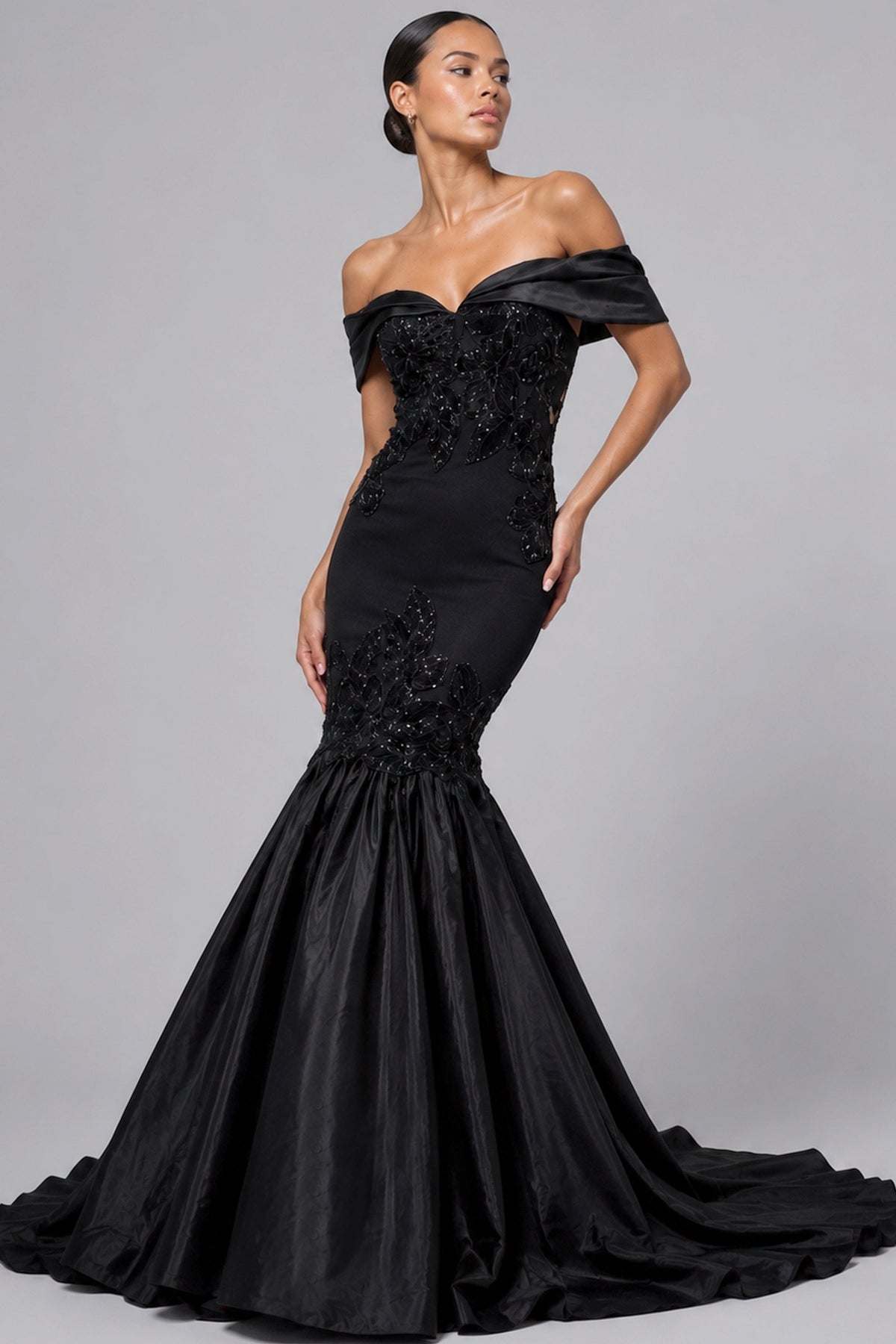 A woman modeling an off-the-shoulder black mermaid gown with a fitted embellished bodice and a full satin flared skirt, photographed against a plain light gray studio background.