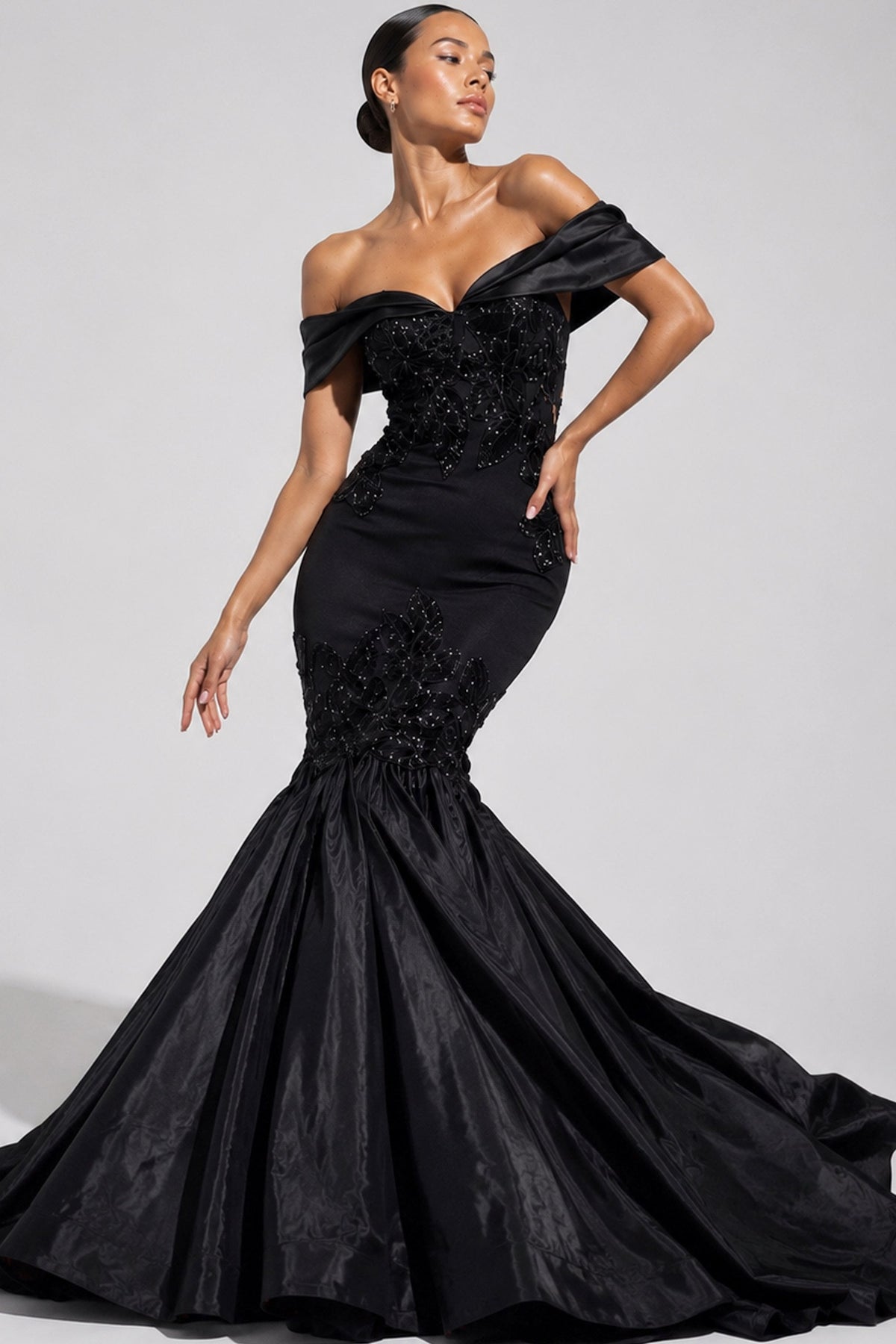 A woman modeling a black off-the-shoulder mermaid gown with a heavily embellished beaded bodice and a full satin skirt, photographed against a light gray studio background.