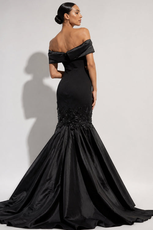 A woman modeling a black off-the-shoulder mermaid gown with a structured bodice, embellished waist, and full satin skirt against a light gray studio background.