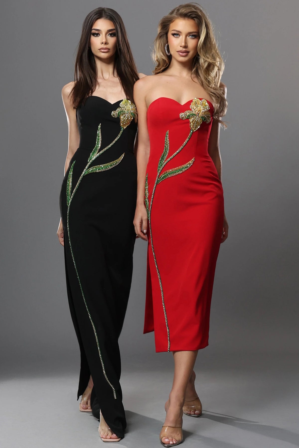 Two models wearing strapless evening dresses with floral and vine embellishments, shown side by side against a gray studio background. One dress is black and the other is red, both featuring gold and green embroidered details.