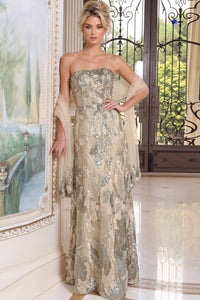 A woman models a strapless floor-length evening gown with shimmering metallic floral patterns in champagne, beige, and silver. She is posed in an elegant interior setting with a large framed painting on the left and a decorative iron gate in the background.