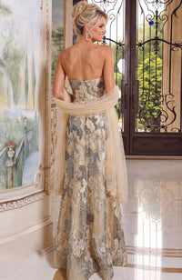 A woman stands indoors wearing a strapless formal gown with a sheer draped overlay. The dress has an abstract floral pattern in beige, taupe, and metallic silver, with a nude back.